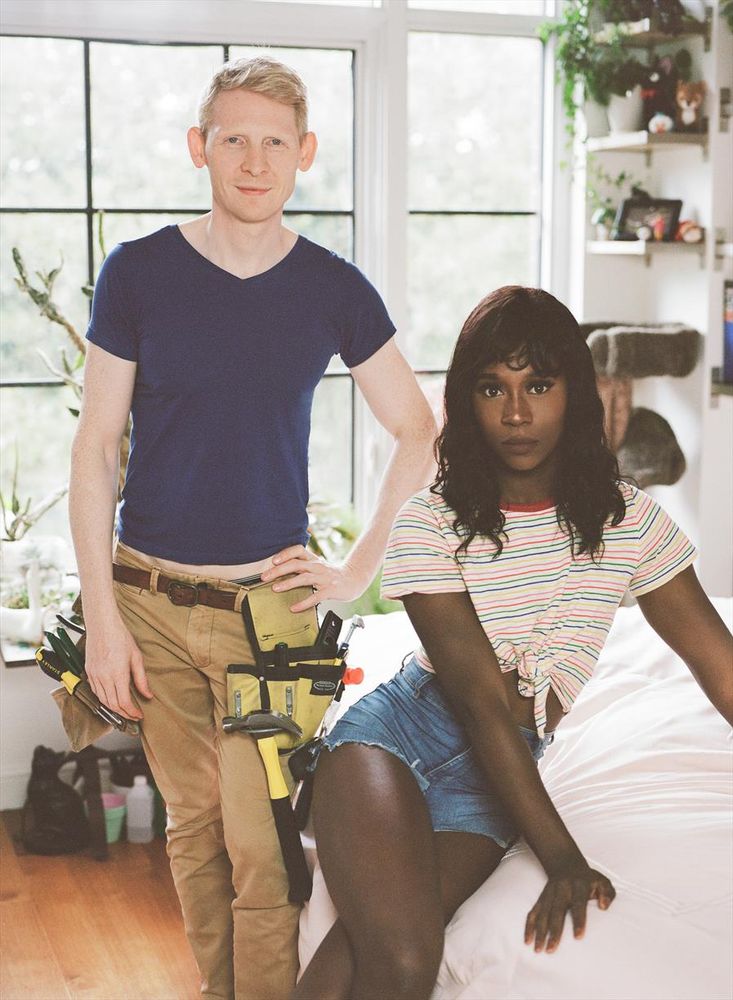 Vanniall sits on a bed wearing a knotted t-shirt and denim cutoffs. Next to her, Rob Yaeger is standing dressed as a handyman. 