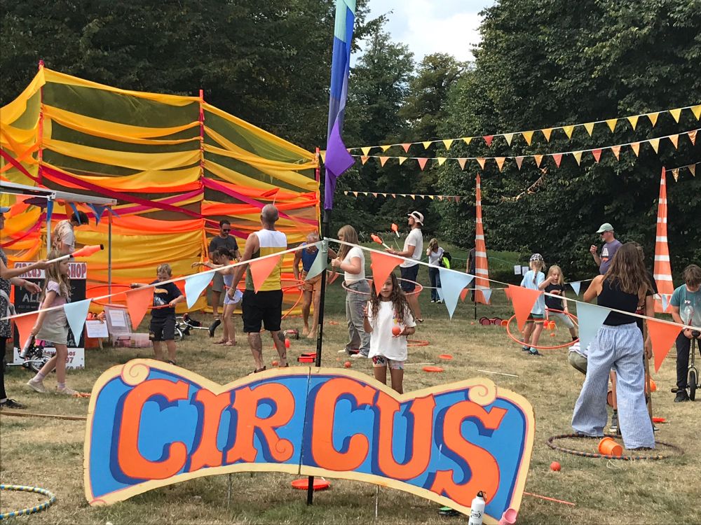 Kids’ areas in The Margins - circus skills like diabolo, juggling and hoops. Photo shows a colourful backdrop of fabric on a screen with a large ‘Circus’ sign, bunting and children and adults having fun playing on the grass