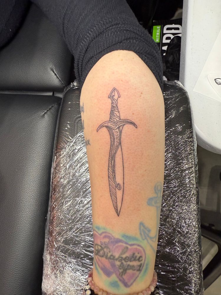 A photo showing a woman’s tattooed arm. She has a sword on her forearm, candy hearts that says Diabetic Type 1 and a blue anchor. Her arm is laying on a black stand with Saran Wrap on it. You can see a black table next to it. There is light shining on her arm. 