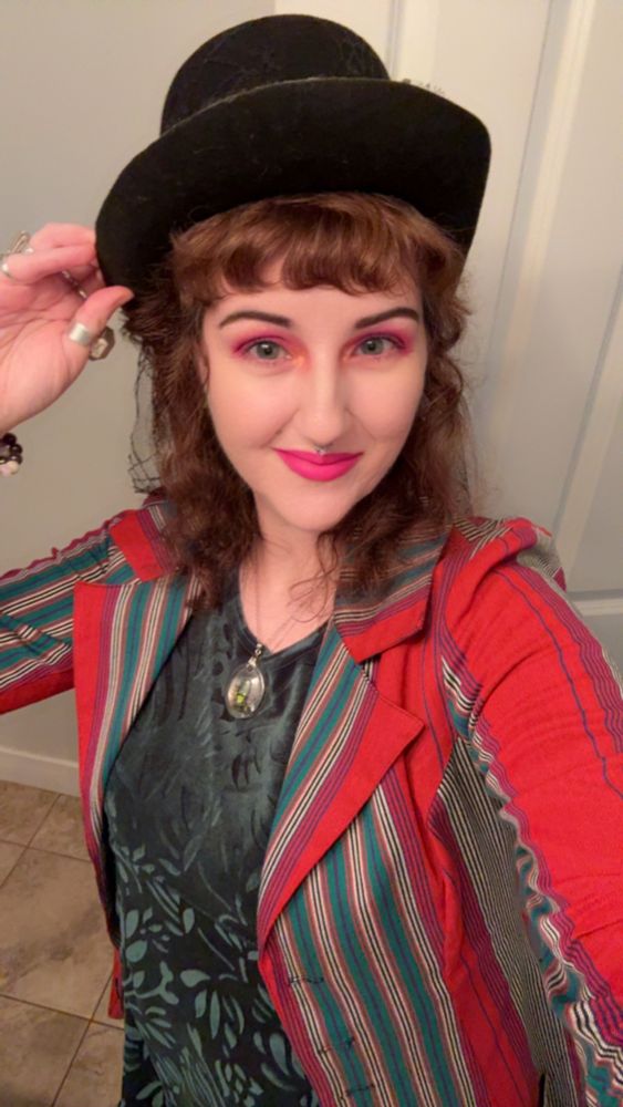 Selfie of me. I’m wearing a striped blazer with red, blue, green and white striped of all sizes, a green velvet dress with floral details and a black top hat with lace. I’m wearing pink eye makeup & pink lipstick and my eyes are blue. My hair is a darker red color. I am smiling. I’m wearing a necklace with a little beetle in it. I’m holding the hat with my left hand. The floor is brown tile. The background behind me is off white. 
