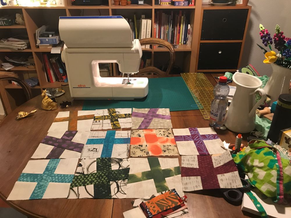 A patchwork in progress. 11 (Swiss! Red?) cross blocks in purple, green, turcoise or orange on greyish background.
#quilts #sewing 