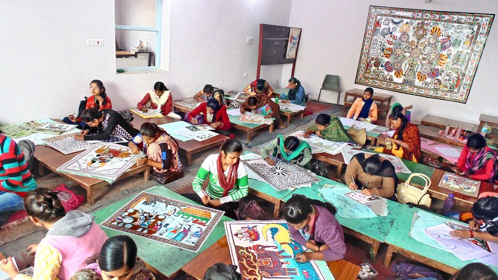 A classroom filled with women seated on the floor at low wooden tables, each focused on creating intricate Mithila paintings. The artists work with fine brushes and pens on colorful designs laid out before them. The room has plain white walls, and a large, elaborate Mithila painting is prominently displayed on the back wall. The atmosphere is vibrant and industrious, reflecting a collective dedication to traditional Indian art forms. Some women wear scarves and shawls, adding to the colorful and lively scene.