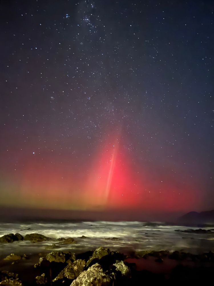 Distinct Aurora rays extending up from the horizon in a starry sky  with waves breaking on rocks in the foreground. 