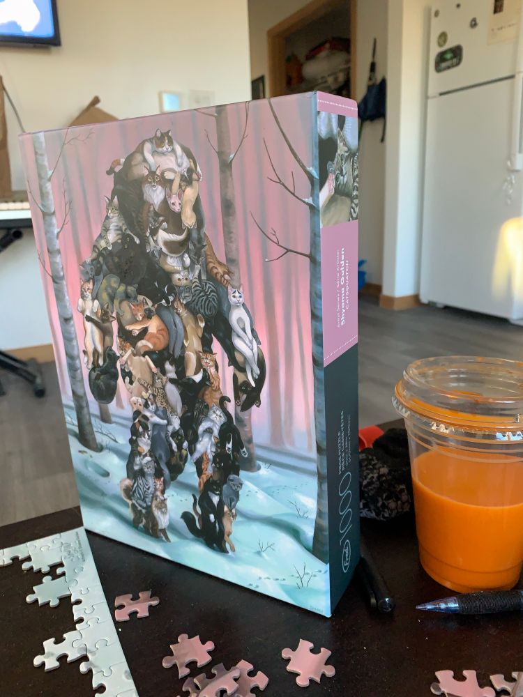 the top of the puzzle box, propped up. it displays a softly lit forest, the subject being a large form on two legs. the form is constructed of many cute kitties 🐱