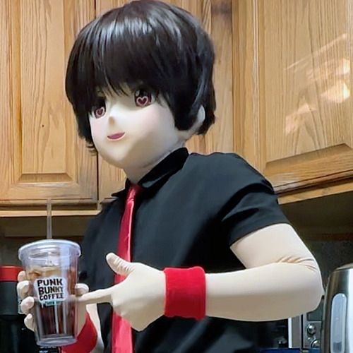 It's me! i'm Billie, a kigurumi character. I'm wearing a black short-sleeved shirt, red tie, and red wristbands. I'm in a kitchen. I have a plastic tumbler filled with ice coffee in my right hand, while I'm pointing to the logo on the glass (Punk Bunny Coffee) with my other hand