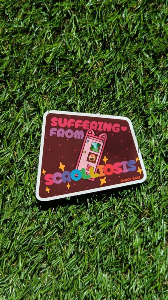 A sticker on a fake grass background. The sticker reads “suffering from scrolliosis” and features an illustrated image of a smartphone with cat ears. 