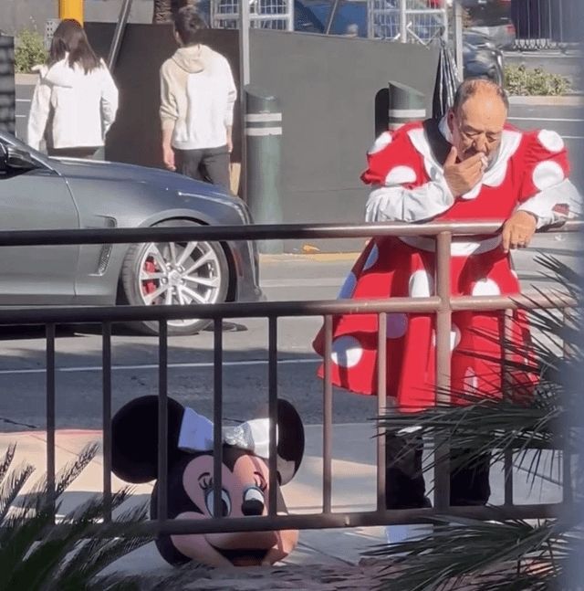 A person is leaning over a railing outside and smoking a cigarette. They're masculine appearing and middle aged. They're dressed in a red and white polka dotted dress with a Minnie Mouse head resting on the ground next to them. The appearance is that they are a Minnie Mouse mascot actor taking a smoke break.