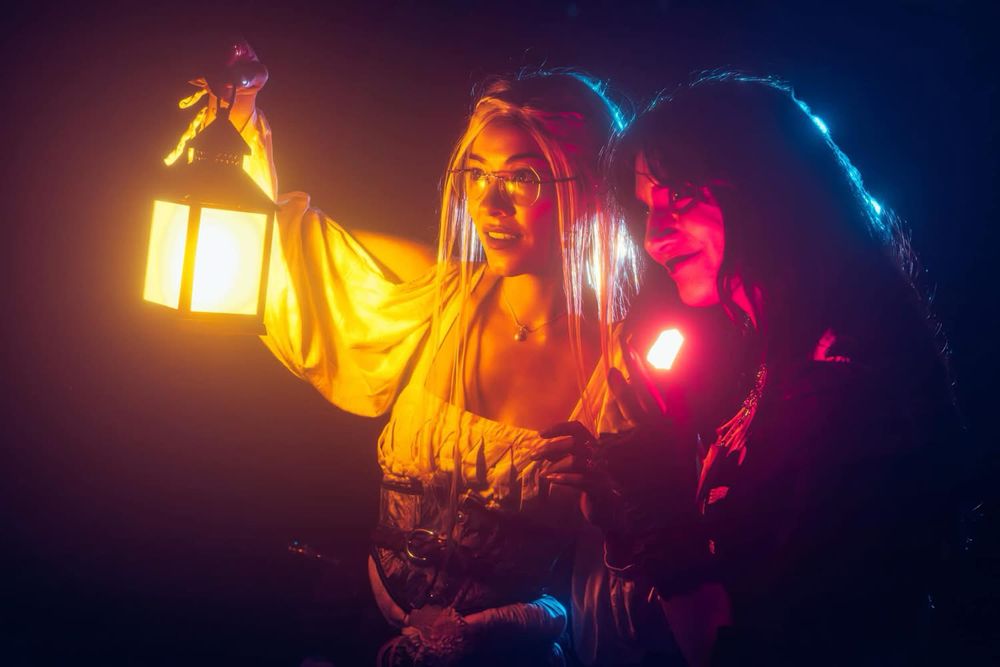 Two femme presenting people dressed in witchy attire. Scene is during night. Both are looking off to the left side of the shot, one in white holding a lantern glowing yellow, the other in black holding a red glowing crystal.