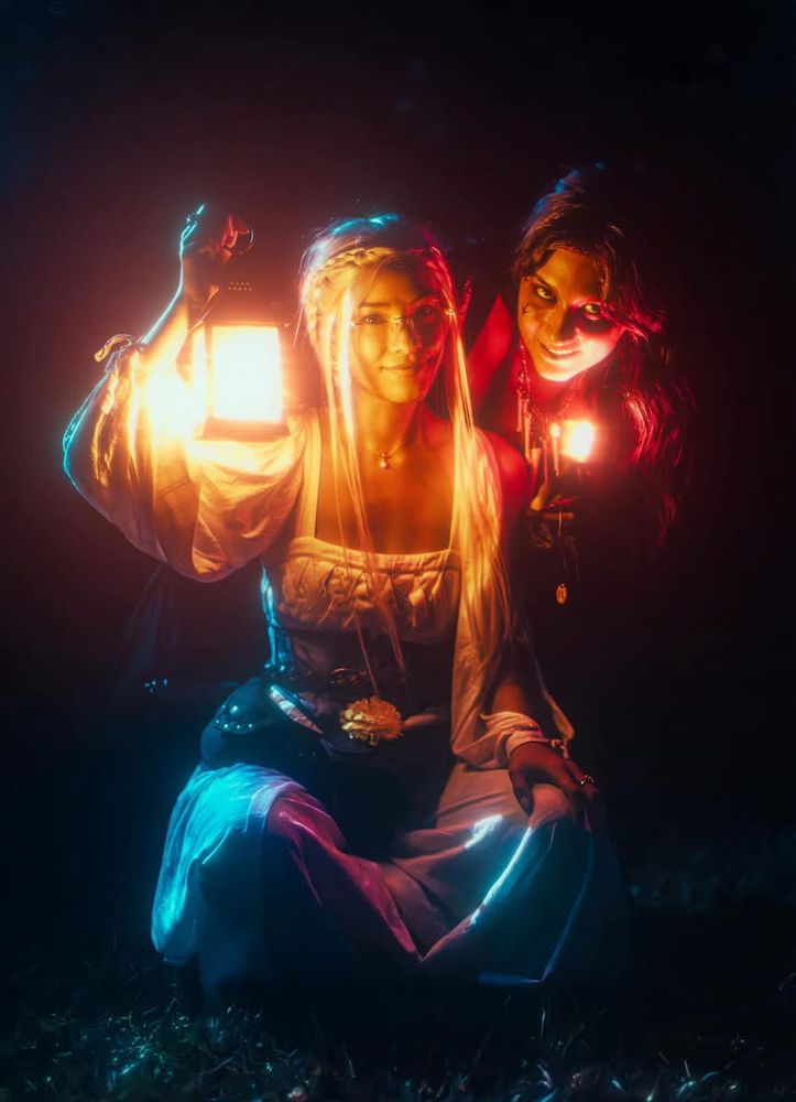 Two femme presenting people dressed in witchy attire. Scene is during night. , one in white holding a lantern glowing yellow and sitting cross legged on the ground, the other in black holding a red glowing crystal leaning over the other one’s shoulder 