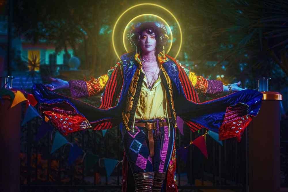 Portrait photo of a Molymauk cosplayer. There’s cloak is bellowing behind them with both hands raised to the side. There’s a glowing halo behind them