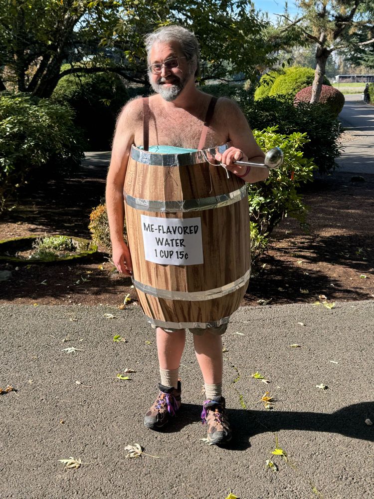 Man wearing costume of a barrel filled either water which he’s soaking in and there’s a sign in the front advertising “Me-flavored water” for 15c a cup.