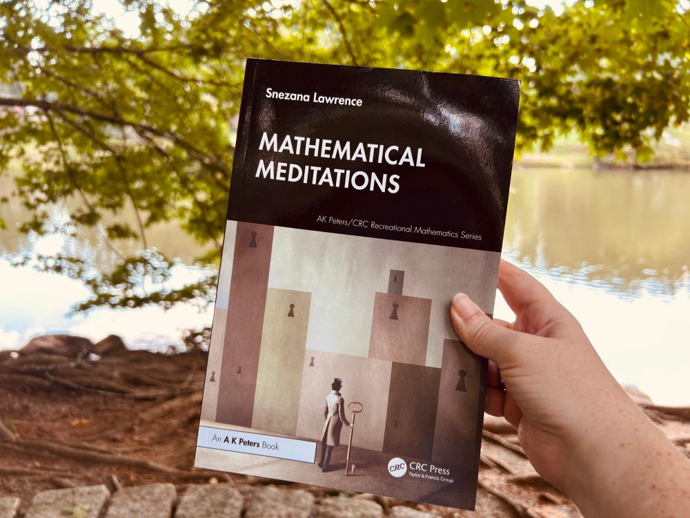 A hand holding Snezana Lawrence’s “Mathematical Meditations” in front of a tree and lake. The cover of shows a building composed of white, green, gray, and brown boxes with keyholes and a person standing in front, holding a key.