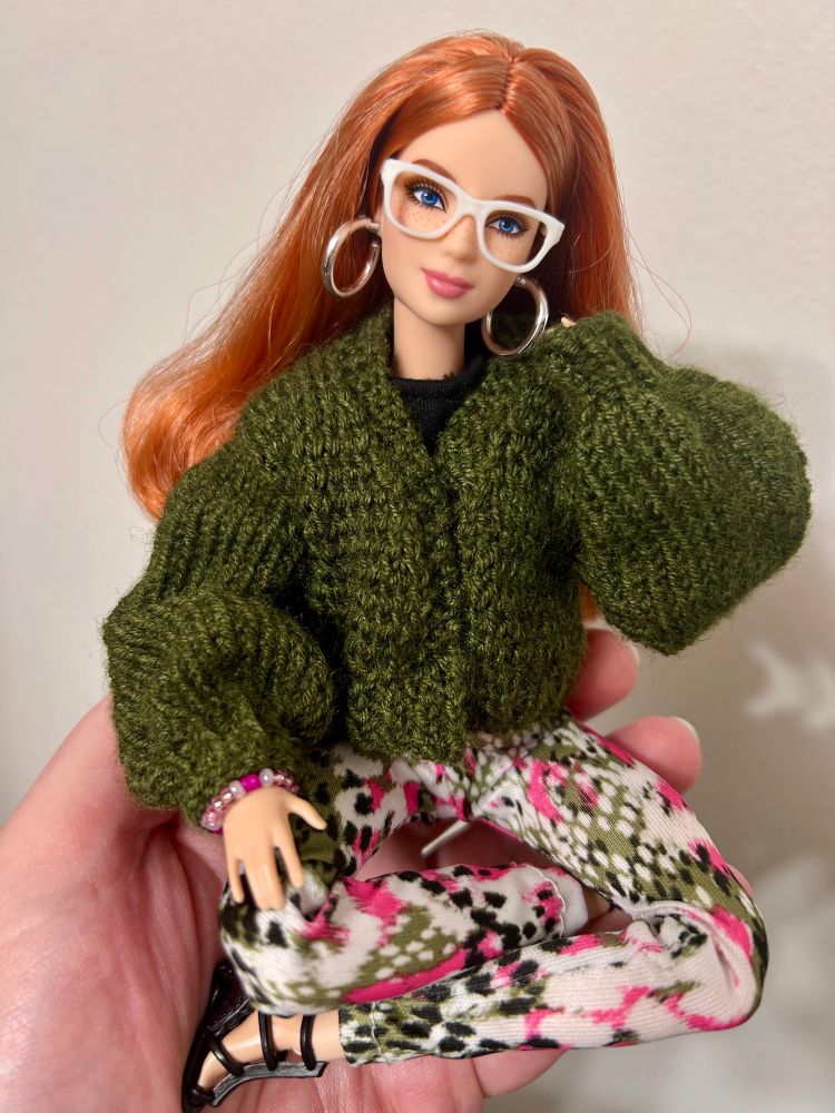 Barbie Basics 2025 Doll 4 sits lotus position in a palm wearing spotted white, pink, black, and olive green leggings, black sandals, a black T-shirt and a handknit olive green balloon sleeved sweater. She also has white glasses and silver hoop earrings.
