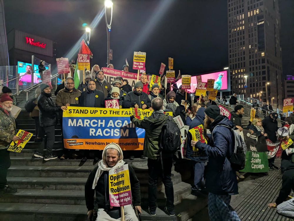 a photo of a large group of protestors holding antiracist placards, banners and flags. the crowd are gathered on steps in Newham in the evening, several organising groups can also clearly be seen. placards show messages such as 'Stand up to the far right', 'march against fascism' and 'stand up to racism' 