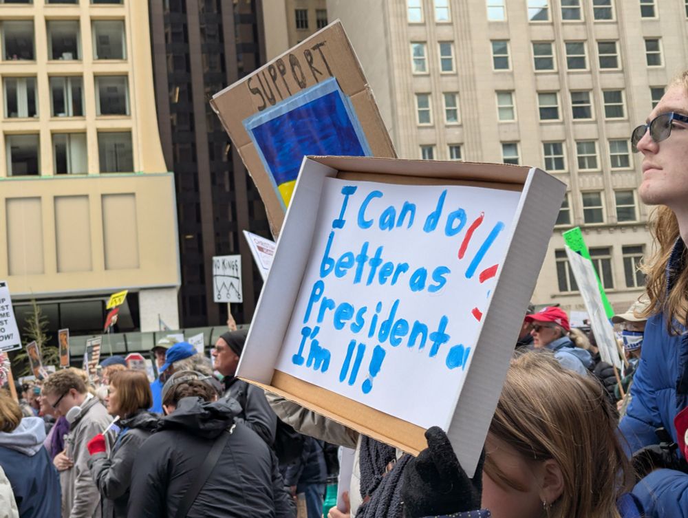 A tenacious young girl (face obscured) carries a sign reading "I can do better as President and I'm 11!"
