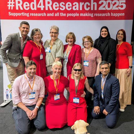 A group of the R&D Forum Trustees and Working Group Chairs and Jessie the dog. Most of the group are wearing red and smiling at the camera in front of a #Red4Research banner.