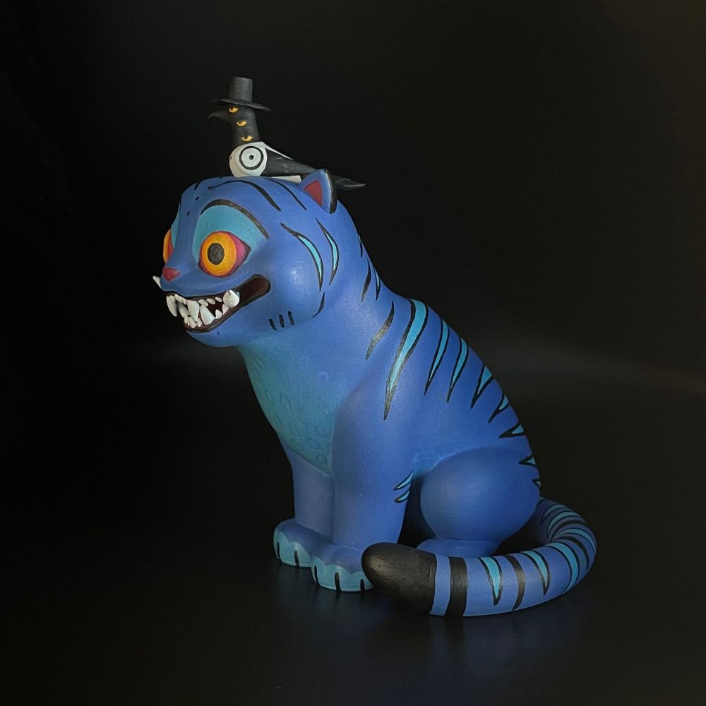 The tiger and bird from kpop demon hunters sitting. It’s a 3d print that I painted. The tiger is blue with red and yellow eyes and a big toothy grin, on its head sits a 3 eyed magpie with a little black hat. 