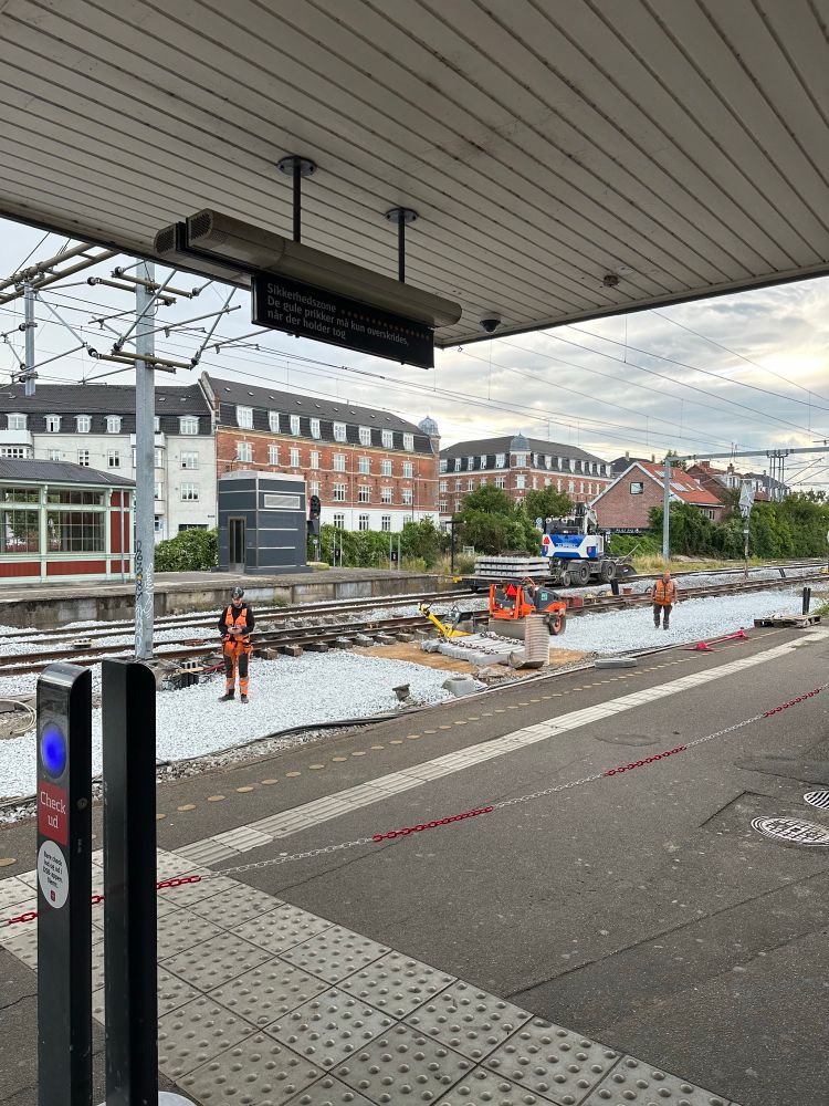 Small construction site at an active commuter rail station. A few workers separated from rest of station with a small chain. They have materials and a few very small construction vehicles. Work was happening. 