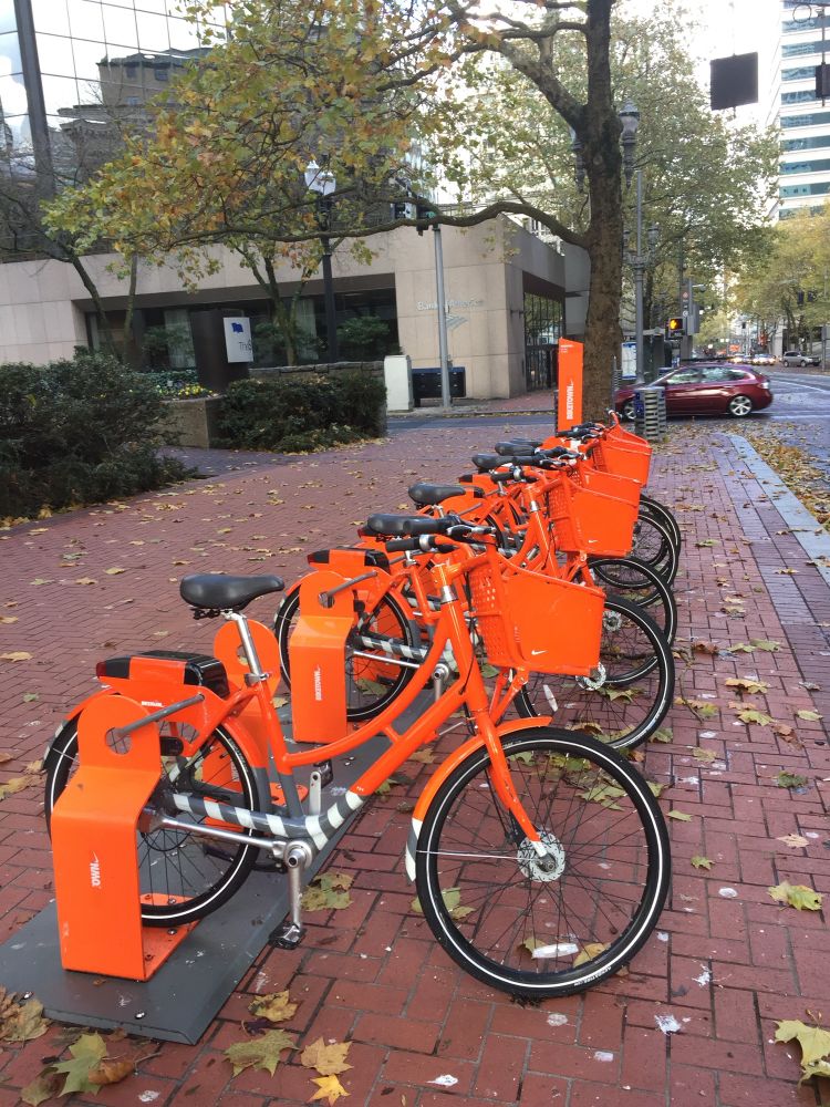 A rack of orange biketown bikes in Portland Oregon. On brick sidewalk with leaves on the ground. 