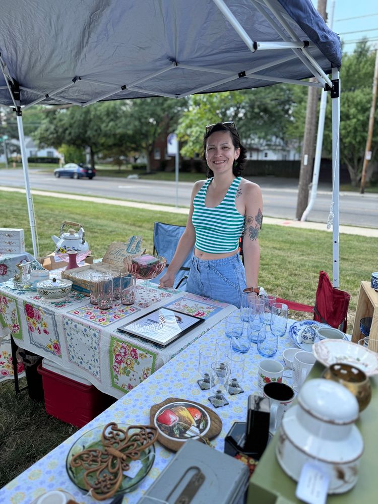 A brunette woman stands behind an "L" shaped table with a tent canopy overhead. The tables are covered in vintage tablecloths and feature a display of vintage glassware, trinkets, and decor.