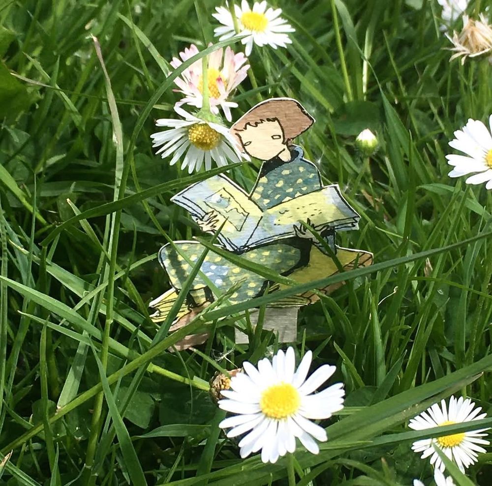 Personnage en papier probablement créé par Martine Delerm, une femme assise avec un livre ouvert dans les bras, cette femme est assise dans l'herbe bien verte parsemée de fleurs, des marguerites