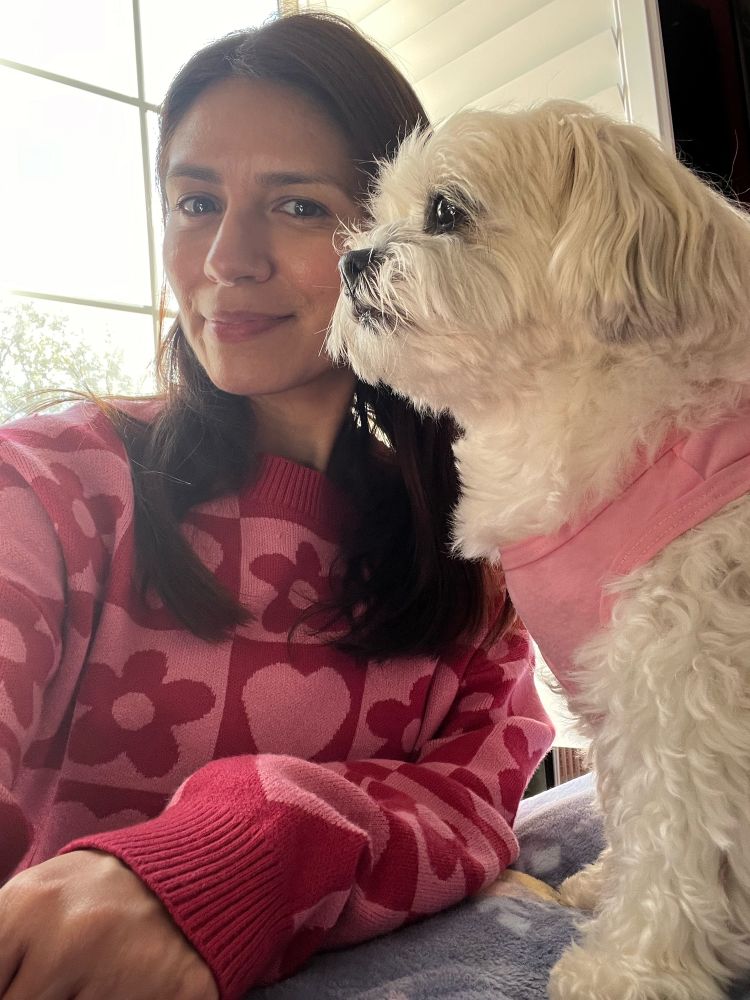 Ashley, smiling at the camera, in a magenta floral sweater. Her dog Emme taking up half the shot. 