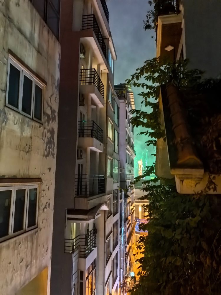 A picture of green and orange fireworks in the distance situated between building complexes in Ha Noi.