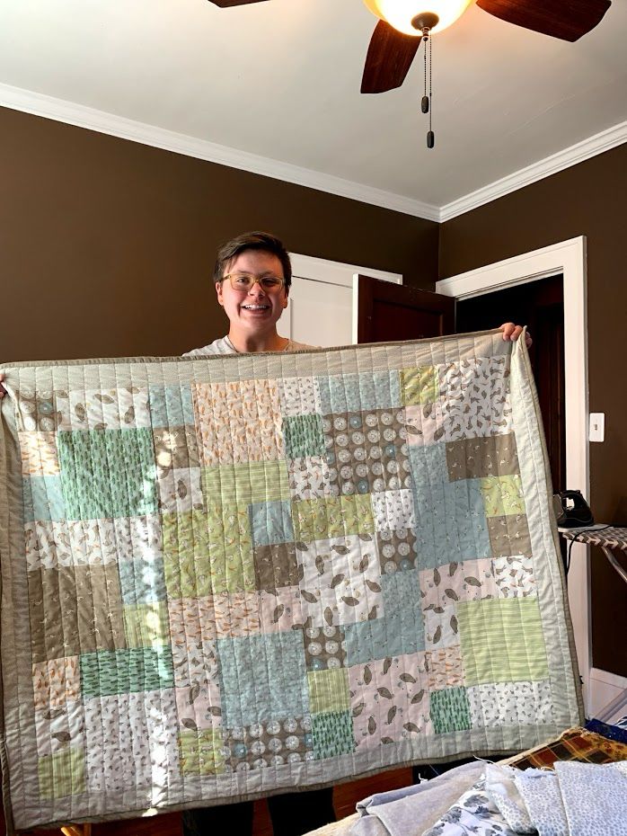 Photo of person holding up a baby size quilt that has neutral colors of green, browns, white and little animals. 