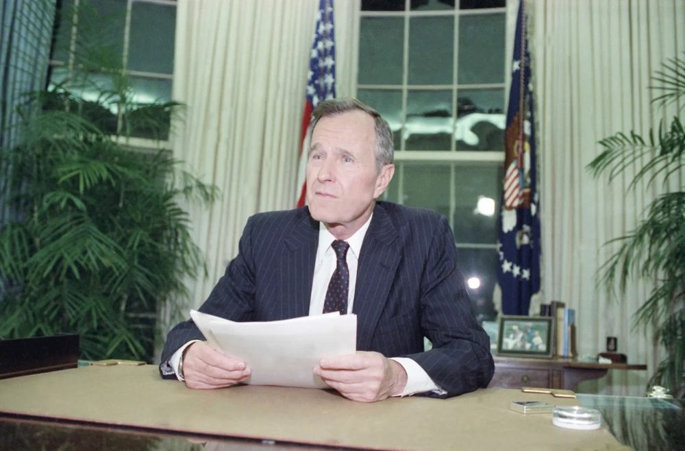 George HW Bush in the Oval Office, wearing a blue coat and tie, sitting at a desk, holding a few papers as he addresses the nation.