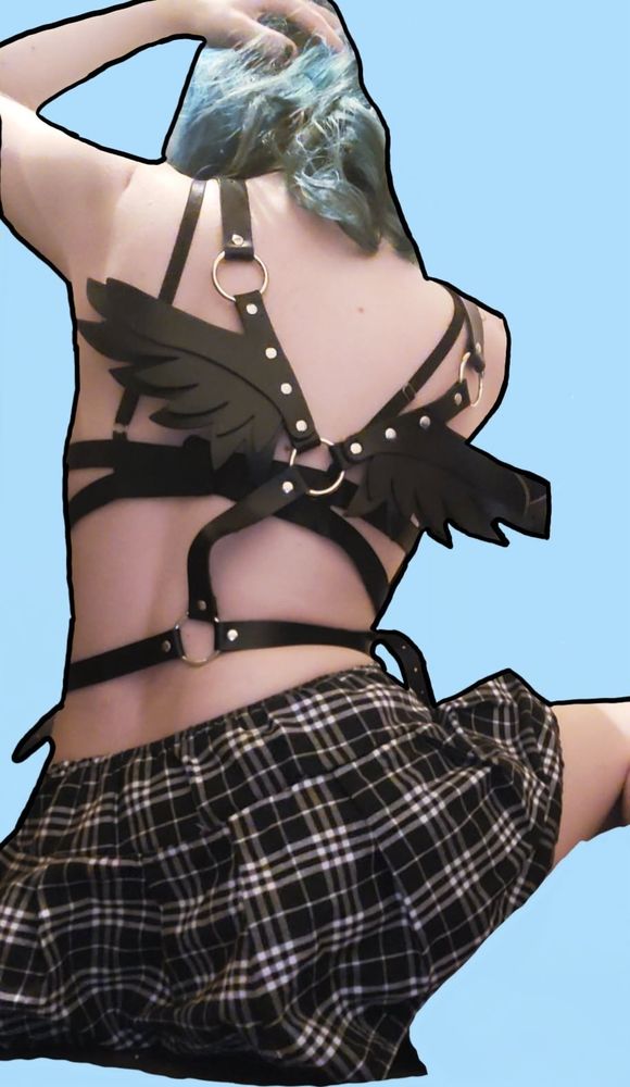 Vampiricc, sitting down on the floor. She is wearing a black bra, a black harness that has angel wings, and a white/black plaid miniskirt. She is facing away from the camera, with her hand in her hair. Her hair is faded blue/aqua. The background is baby blue with a black outline around her body. 