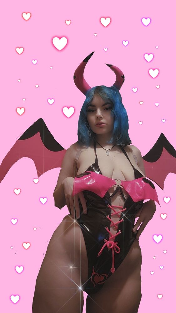 Vampiricc, standing up. She is wearing moeflavor's helluva boss verosika bodysuit, with the matching wings, and horns. She is also wearing a white crystal necklace, thick black cat-eye eyeliner, and pink lipgloss. Her hair is electric blue, the background is cotton candy pink, and there is red/pink/purple neon hearts in the background. She is looking at the camera, with her hands at her waist. 
