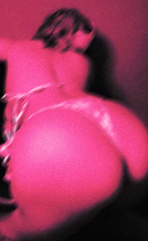 Vampiricc, sitting on a black/white office chair, facing the chair. She is wearing moeflavor's plush cow bikini in pink/white, and white headphones. She is showing off her ass is this photo, and there is a neon pink filter over the photo. 