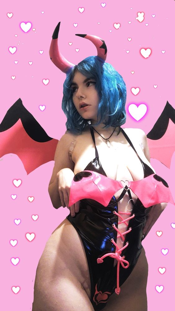 Vampiricc, standing up. She is wearing moeflavor's helluva boss verosika bodysuit, with the matching wings, and horns. She is also wearing a white crystal necklace, thick black cat-eye eyeliner, and pink lipgloss. Her hair is electric blue, the background is cotton candy pink, and there is red/pink/purple neon hearts in the background. She is looking to the side, with her hands at her waist. 