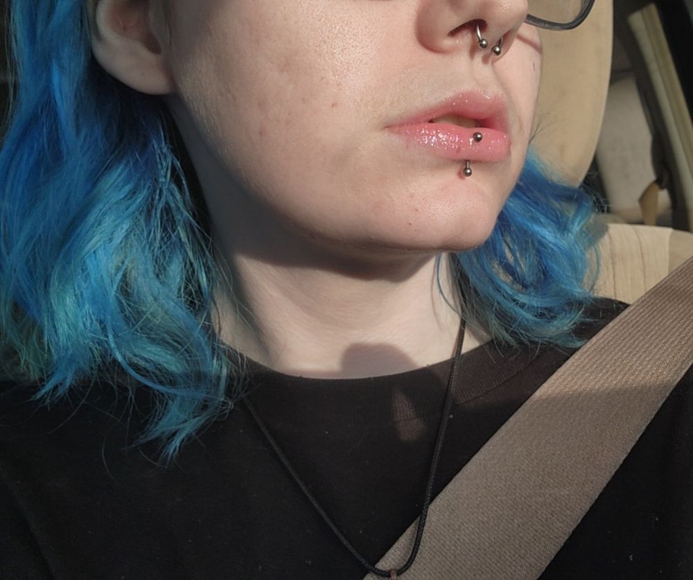 Vampiricc, taking a selfie in the car. She is wearing a black t shirt, a black crystal necklace, and turquoise glasses. You see her silver septum ring, and her silver vertical labret piercing. She has on elf's glow reviver shimmer lip oil "candy coded". 