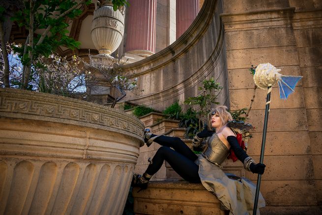 A cosplayer in a black and gold bodysuit and skirt poses magnificently in a classical setting