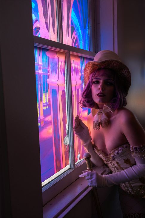 A cosplayer in a heavily decorated white corset, top hat, and gloves poses with dramatic purple lighting in front of a window.