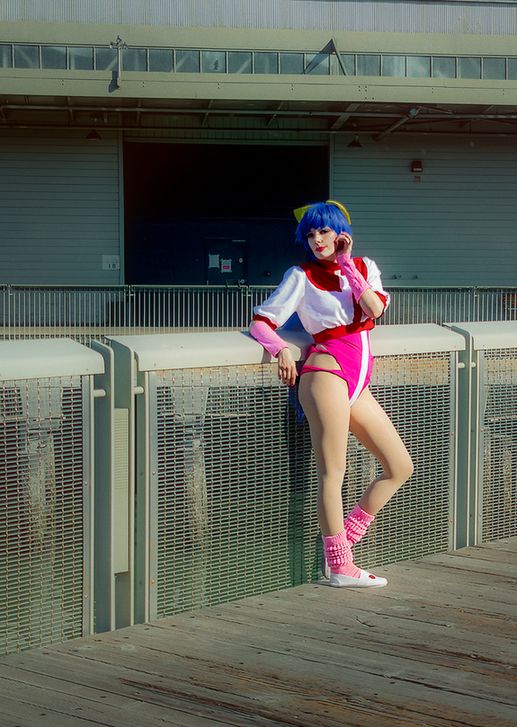 A cosplayer in a blue wig and pink leotard poses at some docks