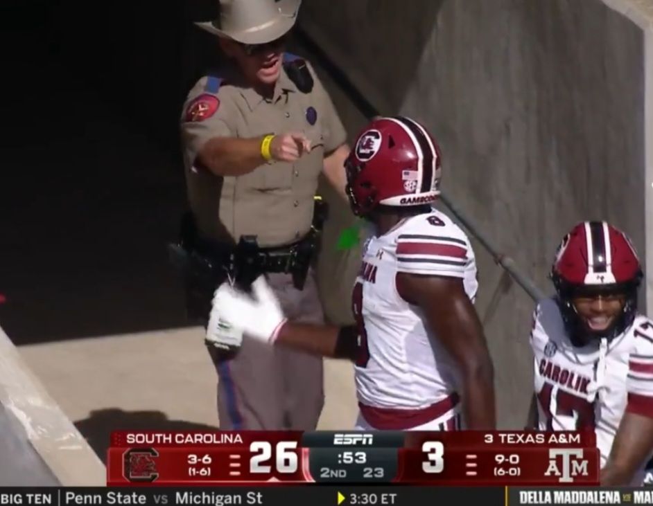 A Texas state trooper in dark shades and cowboy hat points snd shouts at a Black football player after drliberately bumping into him in the tunnel leading to the field.