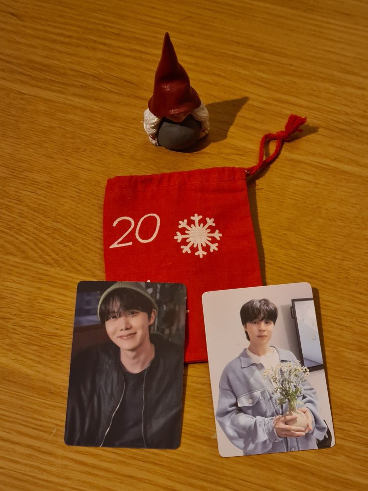 a small cloth drawstring bag with a "20" on it, surrounded by a swedish christmas troll figurine and 2 lomo cards, 1 of hobi wearing a black jacket and green beanie and 1 of jimin in a gray jacket and holding flowers