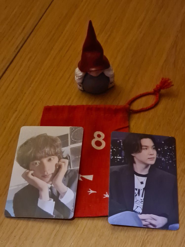 a small cloth drawstring bag with an "8" on it, surrounded by a swedish christmas troll figurine and 2 lomo cards, 1 of seokjin in a suit and tie,  holding his hands to his face, and 1 of yoongi on jimmy fallon, wearing a black blazer and white t-shirt