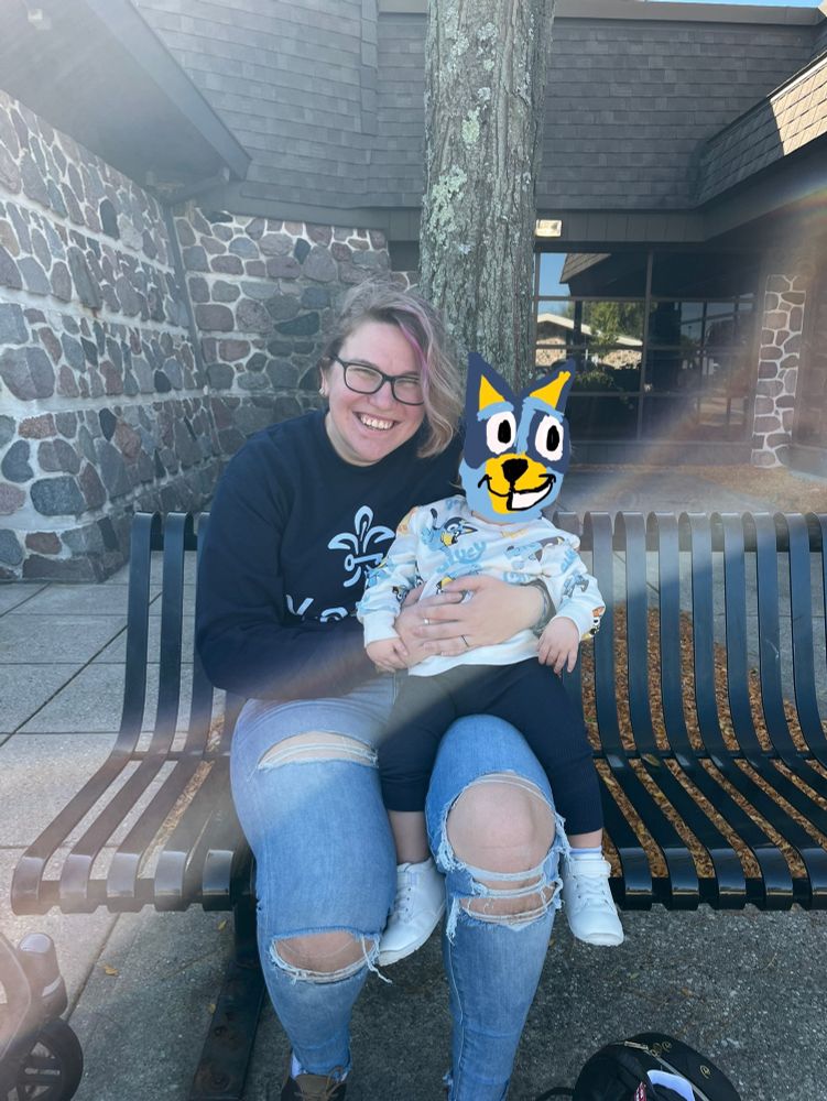 An image of a non-binary person holding their child in a navy blue sweatshirt with light blue writing. They are sitting on a black bench in light wash jeans with holes in them. The child has their face covered by a doodle of Bluey. The child is also wearing a white sweatshirt with Bluey and Bingo printed on it. The child is wearing white sneakers and navy blue pants. Both are smiling outside of the local library - a building made out of grey, brown, and dark colored stone.