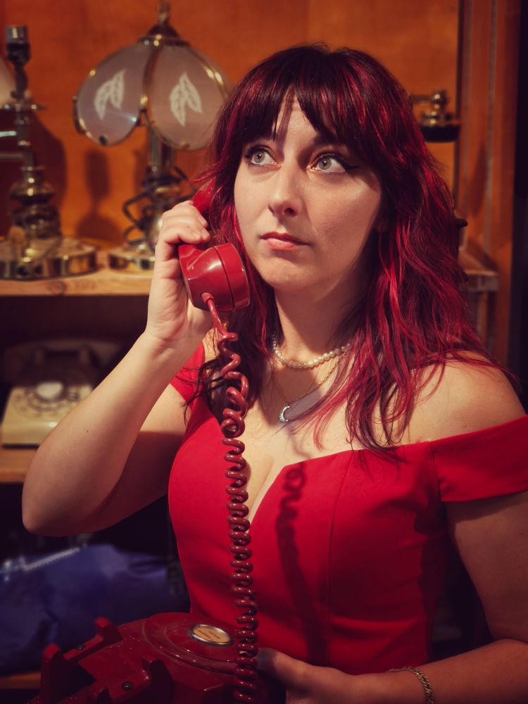 A photo of me edited to look vintage of me wearing a bright red off the shoulder dress with red hair holding a red phone looking off into the distance like a sears photo. I'm all dolled up with makeup and jewelry and standing in front of a shelf of lamps and more phones. 