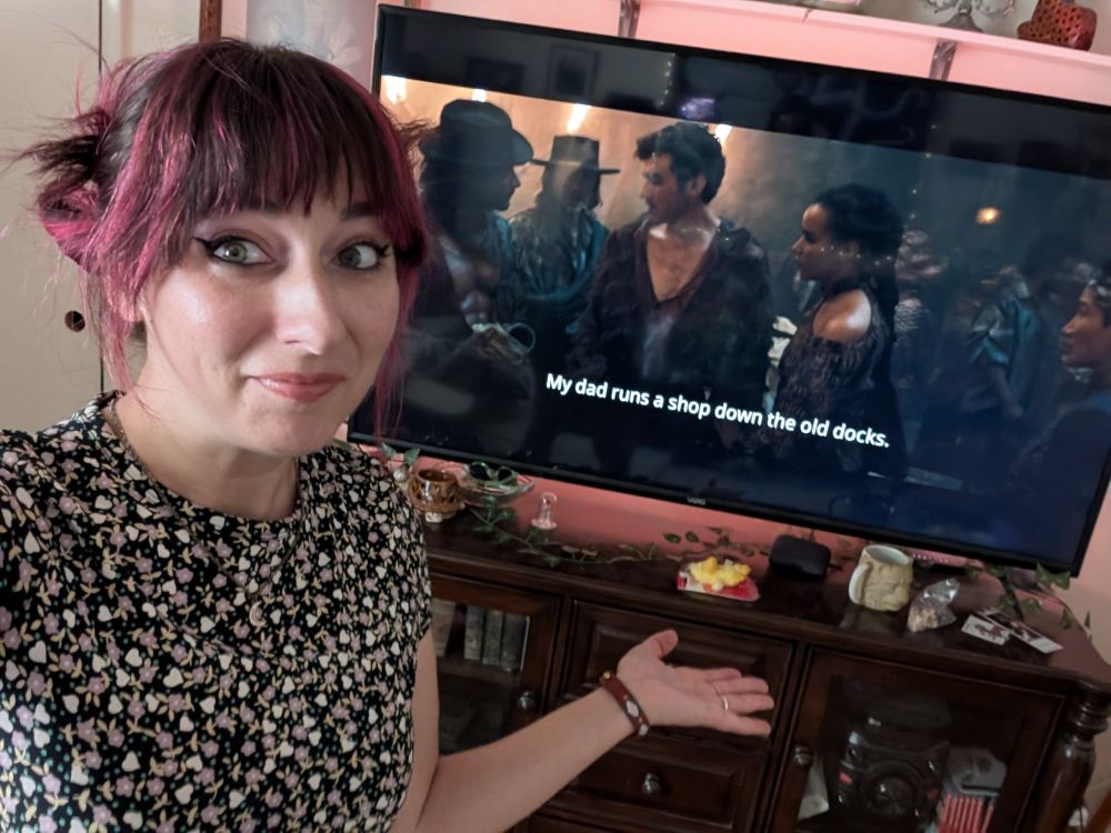 A photo of me standing in front of a large tv with the wheel of time season 3 playing on it. They're in Tanchico. 
