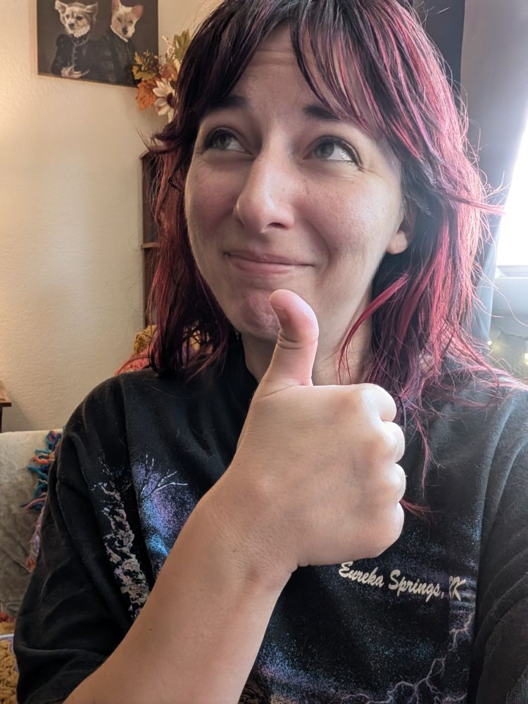 A selfie of me giving a thumbs up and looking up. You can kind of see that I was crying. My hair is red and brown and I'm wearing a black eureka springs tee. 