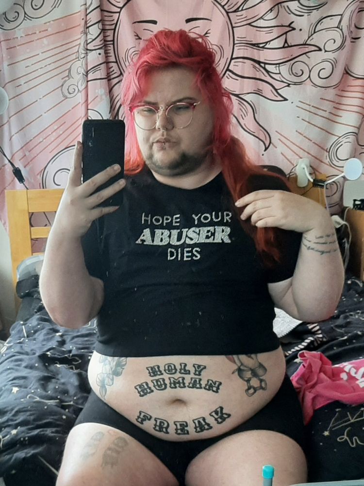 Mx Dagger sits on the end of a bed, their long pink and orange hair coming over their shoulder. They are wearing black boxer briefs and a black crop top that spells out 'Hope your abuser dies' in rhinestones across the chest. Their big round belly is on show, you can read their tattoo that says holy human freak clearly 