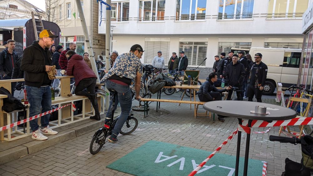 Radfahrer mit Flatterhemd auf Brompton fährt um einen Teppich auf dem "Lava" steht