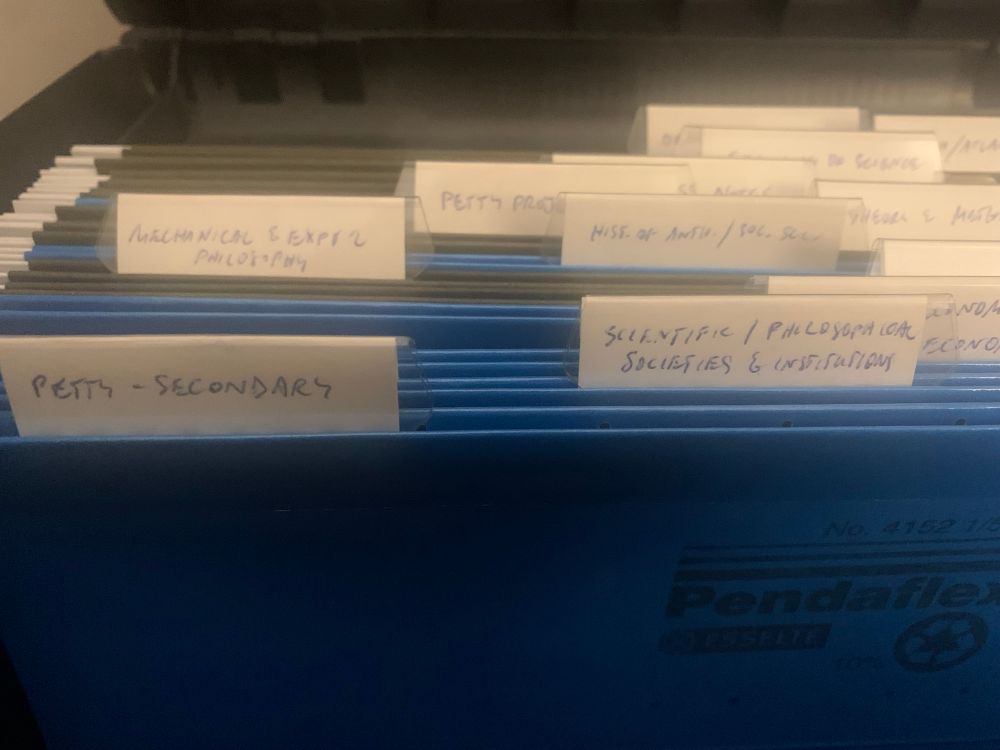 Blue hanging file folders in a box. The folders are empty but have tabs marking the research subject of Ted’s first book. The first tab reads “Petty Secondary,” the second “Scientific/philosophical societies” and so on. 