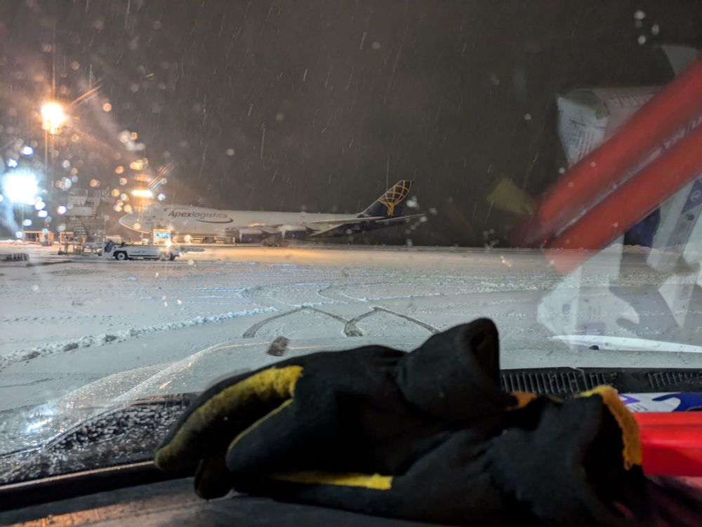 A picture looking out the windshield of a truck. Snow is falling from the sky and the ground is covered in snow, with an airplane in the distance.