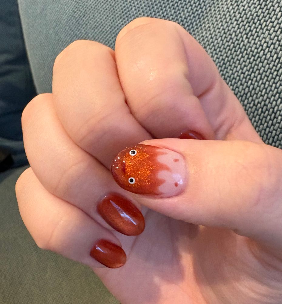 Photograph of right hand. The thumb nail is painted to look like Calcifer from Howl’s Moving Castle with orange magnetic polish. The other four fingernails are painted solid magnetic orange. 