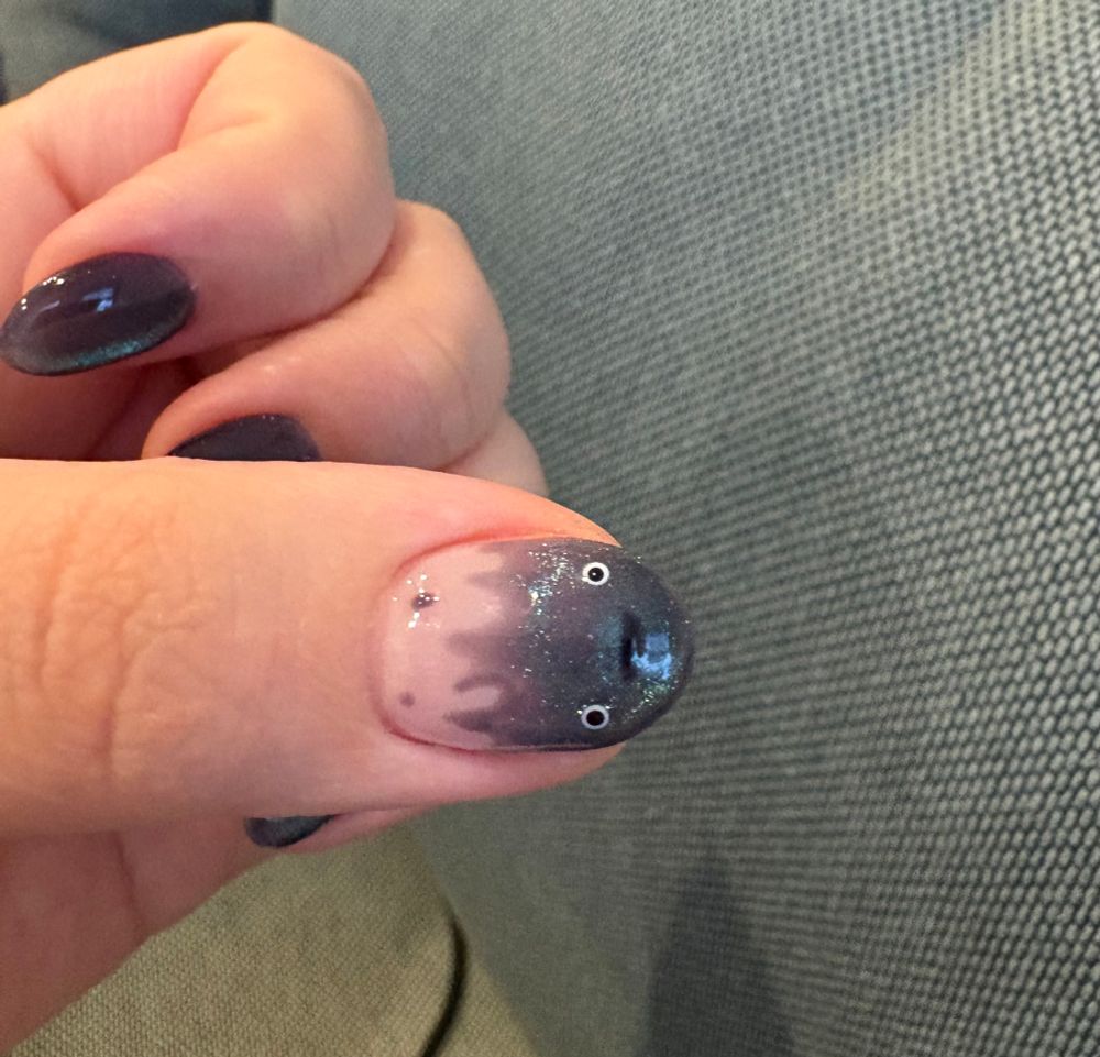Photograph of left hand. The thumb nail is painted to look like Calcifer from Howl’s Moving Castle with blue magnetic polish. The other four fingernails are painted solid magnetic blue. 